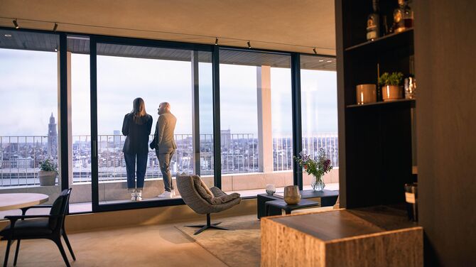 Alt : Vue intérieure de l’appartement moderne avec de larges portes coulissantes et deux personnes sur un balcon.