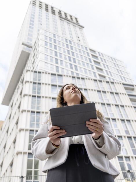 Femme blonde tenant un iPad regardant vers l’immeuble d’appartements blanc.